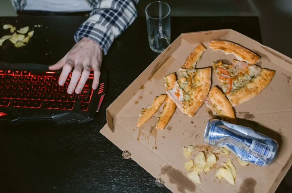 Persona usando el teclado de un pc con sobras de pizza y bebidas azucaradas al lado